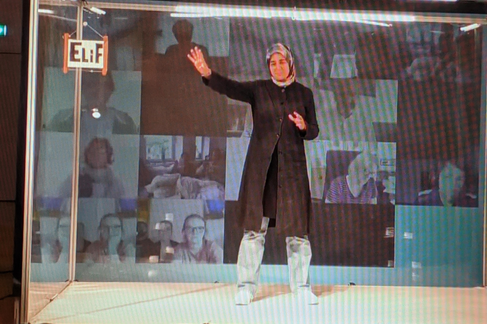 Eine Person mit Kopftuch steht mittig im Bild. Links im Bild ist ein Schild mit der Aufschrift "Elif" zu sehen. Im Hintergrund sind Kacheln einer Videokonferenz eingeblendet.