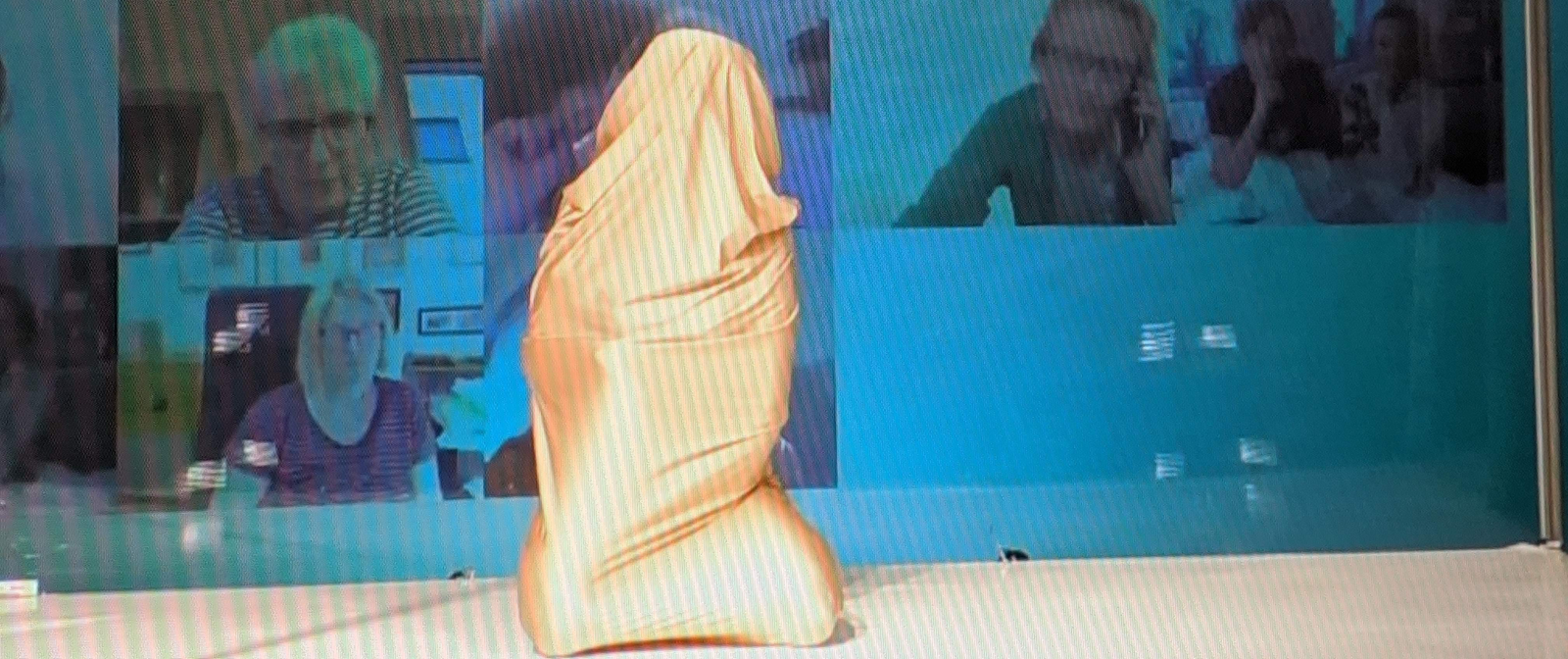 Eine Person ist komplett in ein goldenes Tuch verhüllt und kniet auf der Bühne. Im Hintergrund sind die Kacheln einer Videokonferenz auf einem Bildschirm zu sehen.