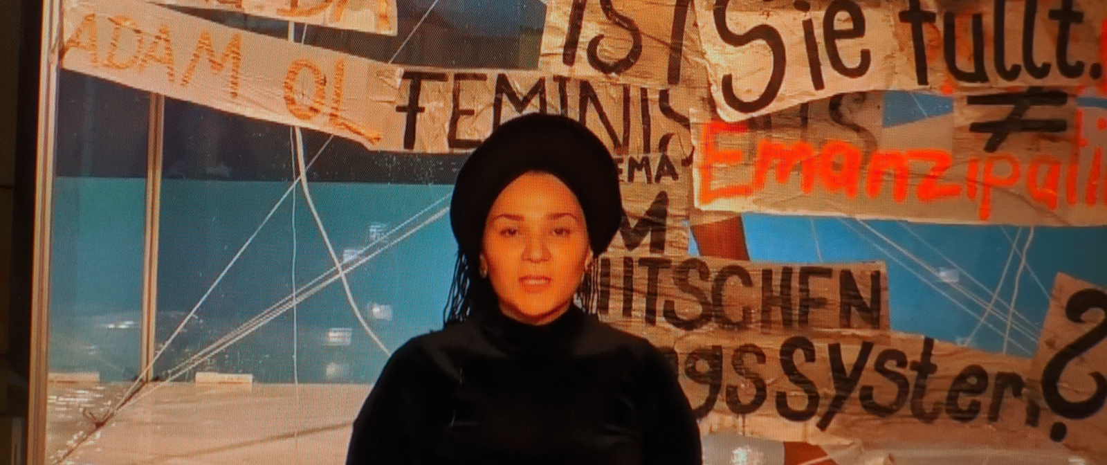 Eine Person im Zentrum des Bildes. Im Hintergrund sind mehrere Banner mit Aufschriften wie "Feminismus" und "Emanzipation" zu sehen.