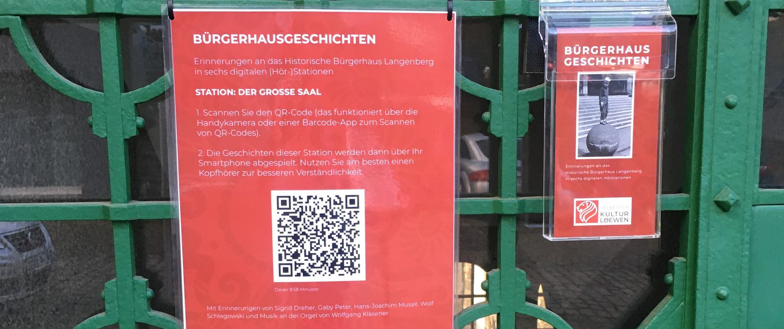 Ein rotes Plakat mit der Aufschrift "Bürgerhausgeschichten. Erinnerung an das historische Bürgerhaus Langenberg in sechs digitalen (Hör-)Stationen. Station 1: Der große Saal". Darunter befindet sich ein QR-Code. Rechts vom Plakat ist eine Acryl-Halterung mit Flyern zum Projekt aufgehangen.