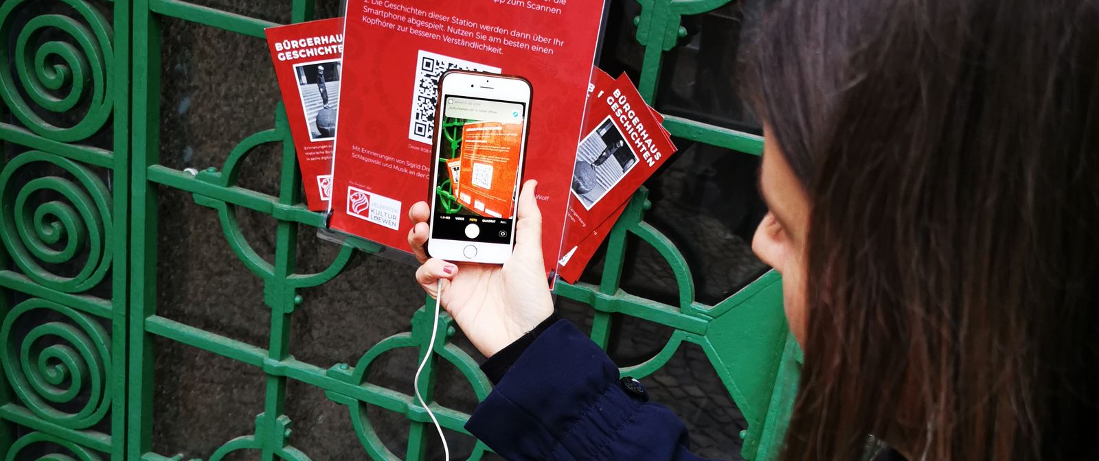 Eine Person vor einem grünen Metaltor scannt mit einem Smartphone einen QR-Code von einem roten Plakat mit der Überschrift "Bürgerhausgeschichten".