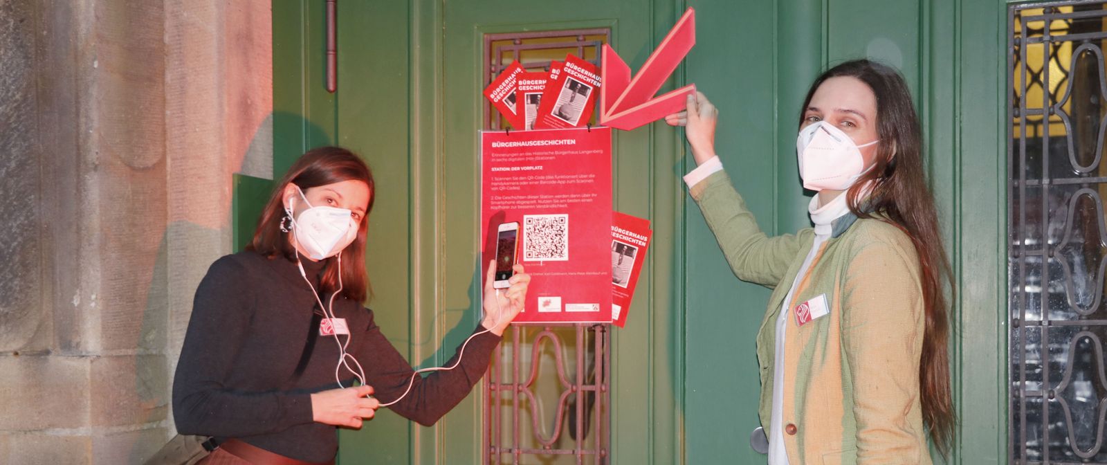 Zwei Personen mit Atemschutzmasken stehen vor einem grünen Tor und zeigen mit einem Smartphone und einem roten Pfeil auf ein rotes Plakat, auf dem ein QR-Code zu sehen ist. 