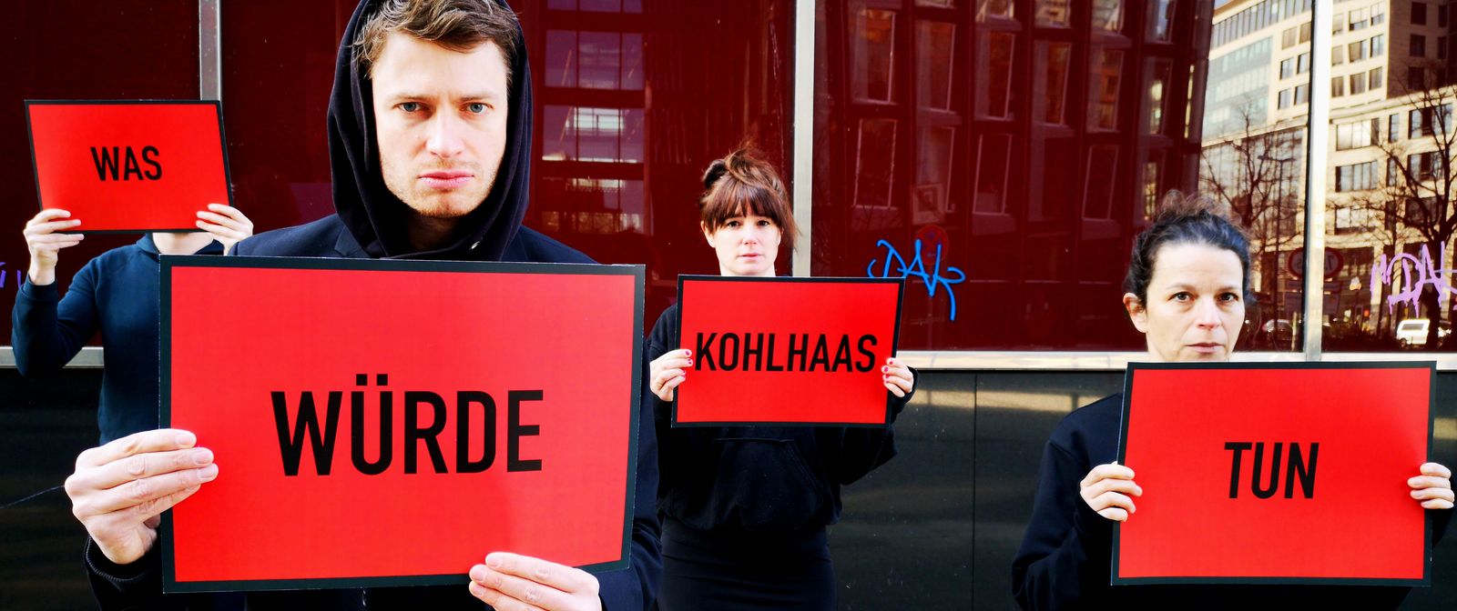 4 Personen im Kapuzenpulli stehen versetzt vor einem Gebäude. Sie halten rote Schilder mit jeweils einem Wort hoch, die den Satz "Was würde Kohlhaas tun" ergeben.