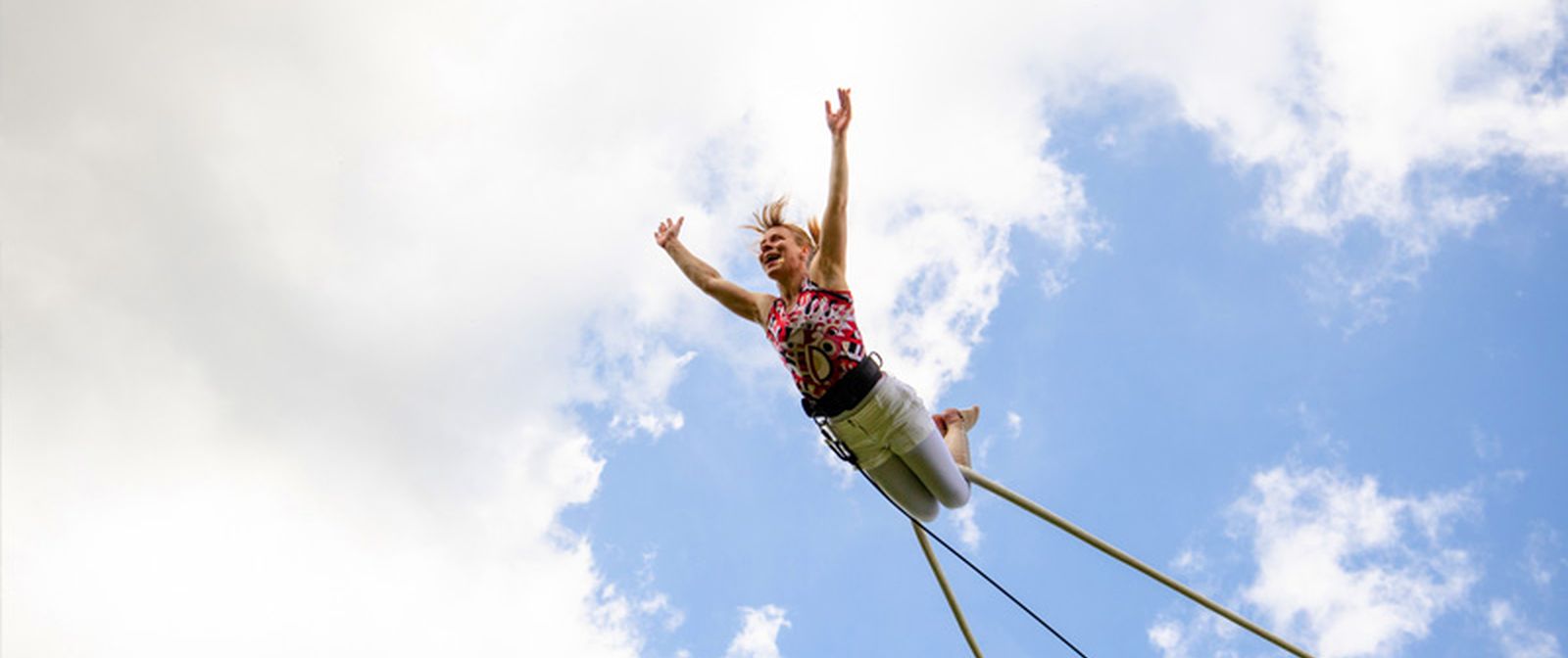 Eine Person hängt mit den Beinen an einer Trapezstange und steckt die Gliedmaßen von sich. Im Hintergrund ist ein blauer Himmel mit Wolken zu sehen. 
