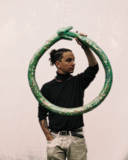Eine Person vor einer hellen Wand. Er hält einen runden Rahmen vor den Körper. Der Rahmen ist eine grüne, glitzernde Schlange.
