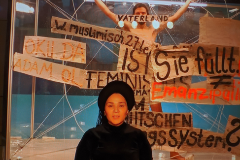 Eine Person im Zentrum des Bildes. Im Hintergrund sind mehrere Banner mit Aufschriften wie "Feminismus" und "Emanzipation" zu sehen. Hinter den Bannern steht eine weitere Person mit erhobenen Händen.