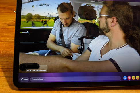 Auf einem Tablet-Bildschirm wird ein Bild von zwei Männern in einem Auto gezeigt. Darunter ist ein Kommentarfeld zu sehen.
