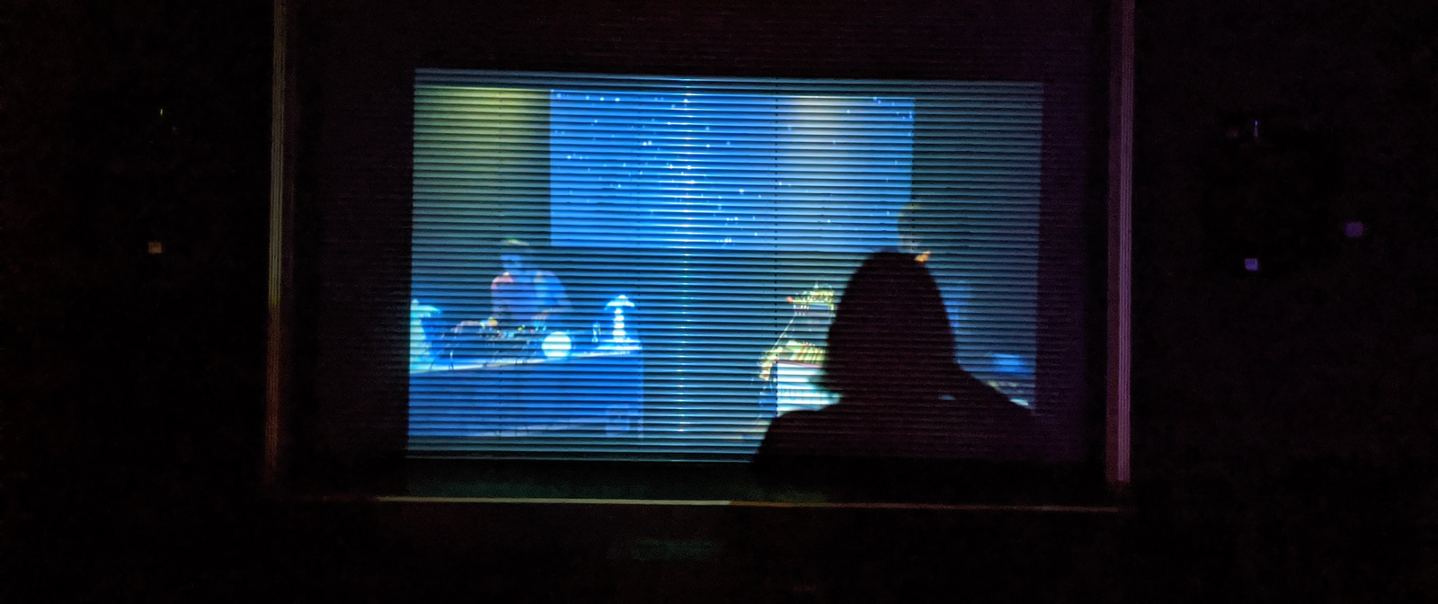 Ein halbdurchlässiges Rollo an einem Fenster lässt ein DJ-Set in dem Raum dahinter erahnen. Vor dem Fenster steht eine Person.