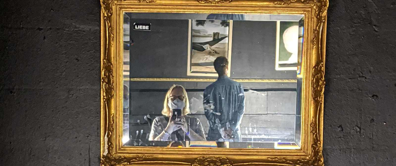 Eine Person mit Atemschutzmaske steht vor einem in einem goldenen Rahmen an eine schwarze Wand gehängten Spiegel und macht ein Foto. Im Spiegel sieht man außer der fotografierenden Person eine weitere, die sich die Gemälde im Hintergrund anschaut.