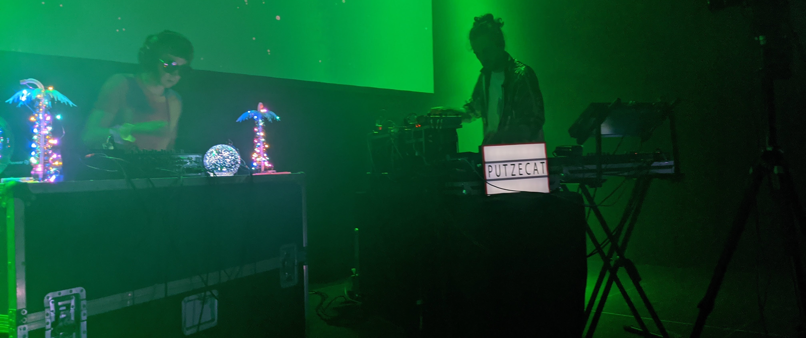 Links sthet ein DJ-Pult mit einer DJ, der die Turntables zwischen zwei leuchtenden Palmen bedient. Rechts ein ein Weiterer Musiker mit einem Keyboard. Auf einem Leuchtschild vor ihm ist der Schriftzug Putzecat zu lesen.