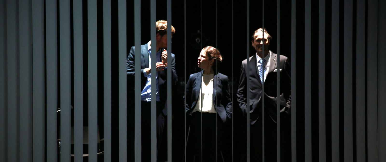 Drei Personen in Anzug stehen in einem dunklen Raum. Die Sicht auf die Personen wird durch einen grauen Lamellen-Vorhang gestört.