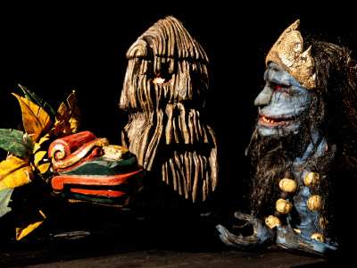 Drei Figuren aus Holz. Links ein bunter Drache, in der Mitte ein holzstammähnliches Gesicht, rechts eine Figur mit blauer Haut und schwarzen Haaren. Die Figuren stehen in einer Gesprächssituation zueinander.  