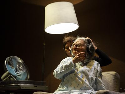 Eine Person hält eine Handpuppe, die das Gesicht einer alten Frau hat, in ein Nachthemd gekleidet ist und Kopfhörer trägt. Der Raum ist durch eine Deckenlampe erleuchtet, links im Bild ist ein Ventilator zu sehen.