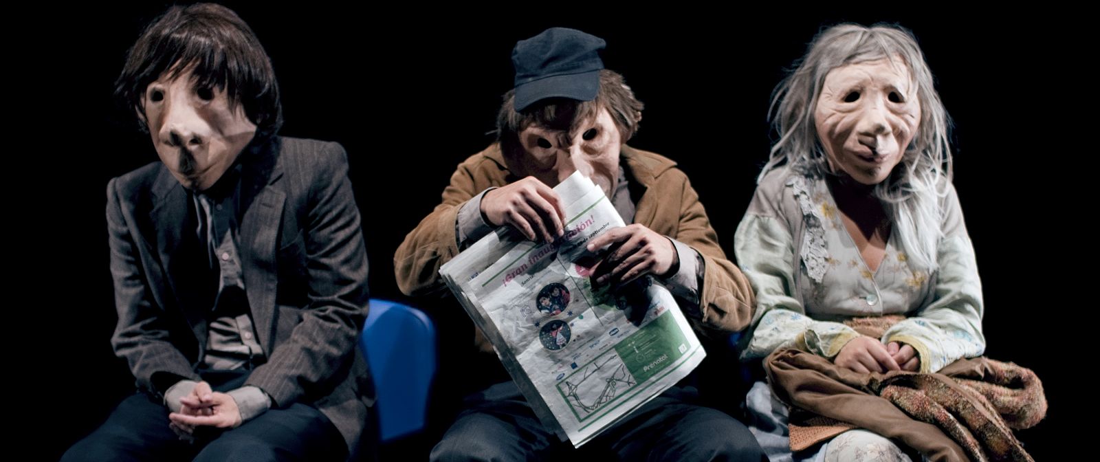 Drei Personen mit menschlichen Masken sitzen nebeneinander und schauen nach vorne. Die Figur in der Mitte hält eine Zeitung in der Hand.