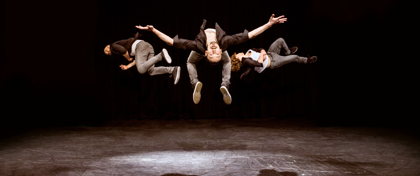 Drei Artisten in dunkler Jeans, weißem Shirt und schwarzem, offenen Hemd springen in die Luft. Ihr Körper sind nahezu horizontal zum Boden.