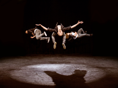 Drei Artisten in dunkler Jeans, weißem Shirt und schwarzem, offenen Hemd springen in die Luft. Ihr Körper sind nahezu horizontal zum Boden.