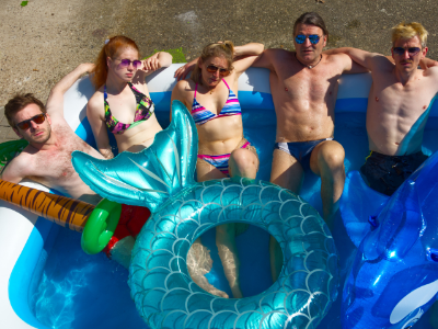 Fünf Personen sitzen in Badekleidung in einem aufblasbaren Pool. Um sie herum sind einige Schwimmtiere zu erkennen. Alle Personen tragen eine Sonnenbrille und schauen nach oben Richtung Kamera.