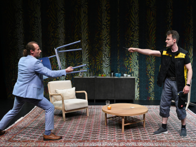 Eine Person in einem blaugrauen Anzug steht links im Bild und richtet einen Stuhl als Waffe auf eine Person, die ihm gegenüber steht und ein Messer auf ihn richtet. Das Bühnenbild zeit einen Sessel, einen Couchtisch, ein Sideboard und einen Teppich. 