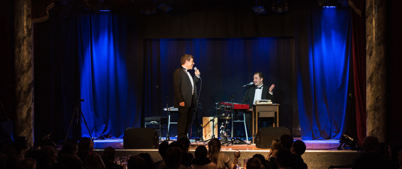 Zwei Männer im Anzug auf einer blau ausgeleuchteten Bühne. Links im Bild steht der Mann mit einem Mikrofon in der Hand, der andere sitzt rechts im Bild an einem Klavier.