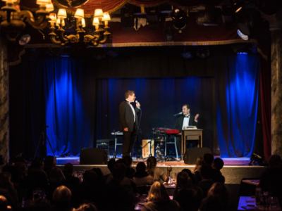 Zwei Männer im Anzug auf einer blau ausgeleuchteten Bühne. Links im Bild steht der Mann mit einem Mikrofon in der Hand, der andere sitzt rechts im Bild an einem Klavier.