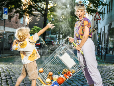 Eine Frau steht mit einem im Umkippen begriffenen Einkaufswagen in einer städtischen Wohngegend. Ein Klienkind spielt mit den Lebensmitteln, die aus dem Einkaufswagen fallen. 