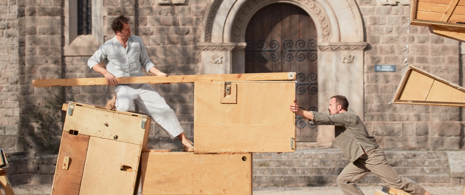 Zwei Männer auf einer Holzkontrustion schieben einen Holzkasten gegeneinader. Im Hintergrund ist alte Fassadenmauer zu sehen.