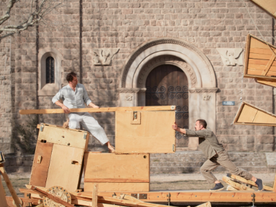 Zwei Männer auf einer Holzkontrustion schieben einen Holzkasten gegeneinader. Im Hintergrund ist alte Fassadenmauer zu sehen.