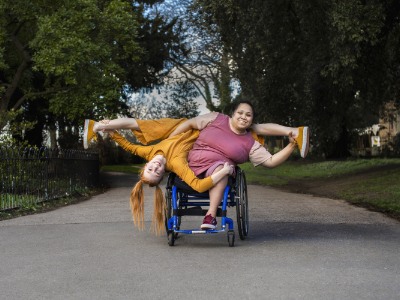 Im Zentrum des Bildes sind zwei Performerinnen zu sehen, von denen eine im Rollstuhl sitzt. Die zweite Performerin lehnt sich von hinten kopfüber und mit den Beinen in einer Grätsche über die sitzende Performerin. 