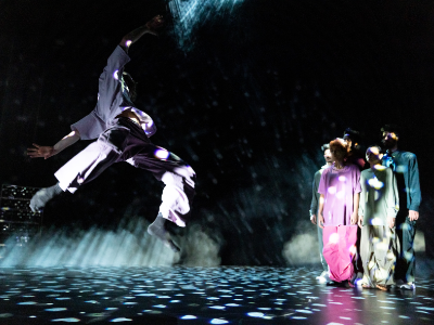Auf der linken Seite der Bühne sieht man fünf Personen in bunten Trainingsanzügen in einer Gruppe stehen. Sie blicken zu dem Tänzer, der sich an der linken Bühnenseite in der Luft zu drehen beginnt. 