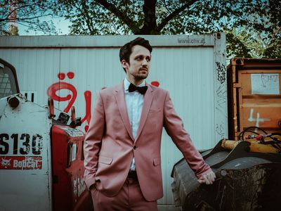Autor Fabian Navarro steht in einem rosafarbenen Anzug vor einem weißen, mit wenigen Graffitti besprühten Übersee-Container.