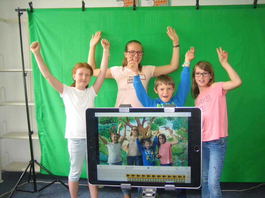 Es ist ein Ipad im Vordergrund zu sehen, welches vier Kinder fotografiert, die vor einem Greenscreen stehen. Die Kinder heben die Hänge und jubeln.