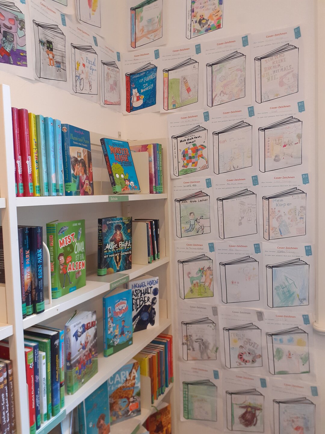 Auf der linken Seite ist ein weißes Bücherregal mit mehreren Kinderbüchern zu sehen. Rechts an der Wand hängen analog gestaltete Buchcover.