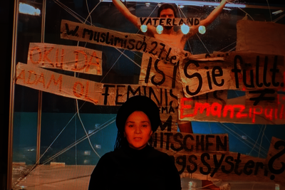 Eine Person im Zentrum des Bildes. Im Hintergrund sind mehrere Banner mit Aufschriften wie "Feminismus" und "Emanzipation" zu sehen. Hinter den Bannern steht eine weitere Person mit erhobenen Händen.