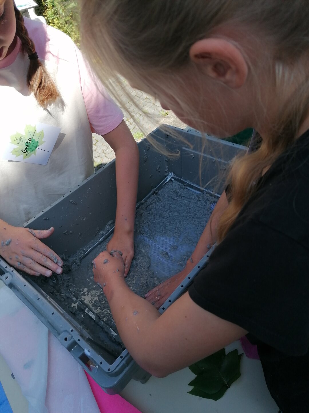 Zwei Kinder schöpfen Papier in einer grauen Plastikwanne.