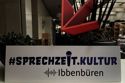 Ein Schild mit der Aufschrift "Sprechzeit.Kultur Ibbenbühren" auf einem Schreibtisch mit Zimmerpflanzen. Oberhalb des Schildes wird ein roter Papppfeil ins Bild gehalten.